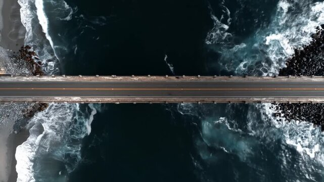 Dynamic aerial drone shot of a coastal road or bridge separating deep blue ocean water and white foamy waves crashing on a dark beach.