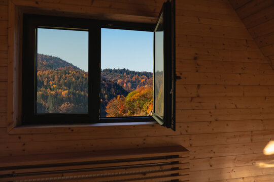 An open window in a wooden cabin looking out at colorful autumn forest