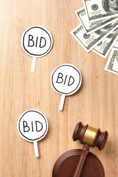 Three white bidding paddles with "BID" signs, auction gavel and US dollar bills on a light wooden surface. Concept of auction, bidding, money, finance and sales. Top view with flat lay.
