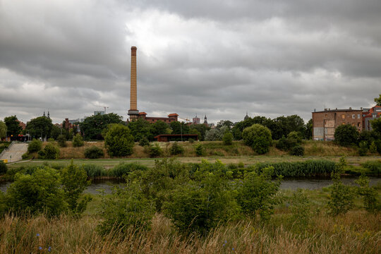 Poznan, Poland - July 8, 2026: Historic gasworks chimney rises beyond the green Warta River floodplain beneath an overcast morning sky.