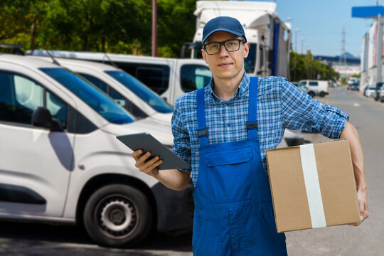 A courier holding a cardboard box and digital tablet with a delivery van in the background.