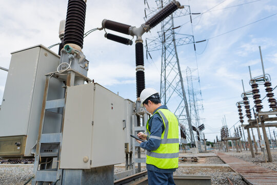 Routine control cabinet inspection at a substation shows an engineer inspecting equipment and writing on a tablet beside a transmission tower.