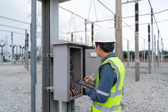 Routine substation cabinet inspection shows a technician using a tablet outdoors at a control cabinet beside a lattice transmission tower.