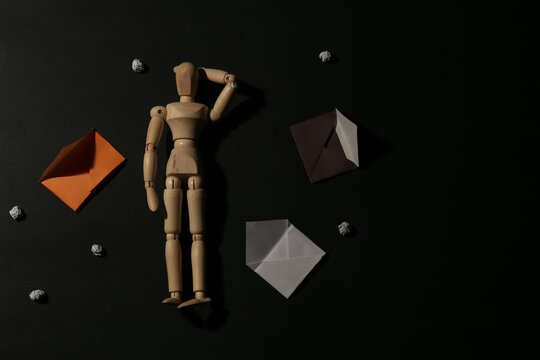 Wooden mannequin looking confused surrounded by empty envelopes and crumpled paper balls on dark black background, concept of confusion, stress, memory loss or bad news.