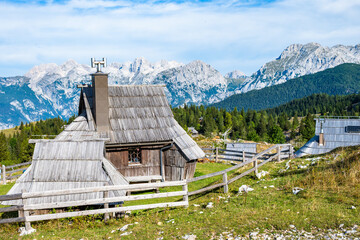 Obraz premium Alpine wooden cottage on Velika Planina set against the dramatic backdrop of the Kamnik-Savinja Alps under a clear blue sky, Slovenia