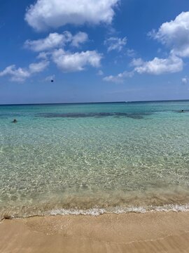 Crystal clear turquoise tropical beach water lapping on golden sandy shore beneath a bright blue sky with fluffy white clouds on a sunny summer day
