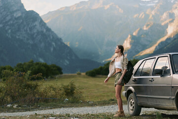 A woman in casual outfit stands beside a car on a dirt road looking at mountain scenery. The photo captures outdoor lifestyle and adventure in nature. © SHOTPRIME STUDIO
