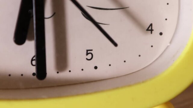 Close cropped analog clock face with white dial, black hands, partial numerals, dot markers, and bright yellow casing, shot from side angle with soft focus background and slight hand blur for