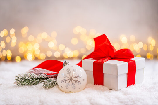 A Christmas background featuring a white gift box tied with a large red satin bow beside a frosted pine branch and red ornament on snow, softly lit by warm twinkling lights with copy space.