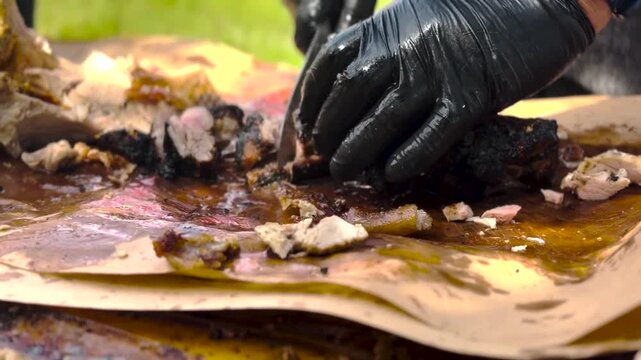 Close-up video footage of a chef wearing black gloves using a sharp knife to slice freshly smoked barbecue meat on butcher paper.
