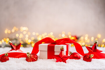 Naklejka premium A Christmas background featuring a white gift box tied with a red satin bow surrounded by red star and bauble ornaments on snow, softly lit by warm twinkling lights with copy space.