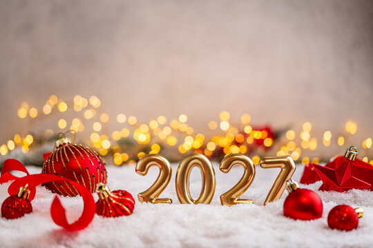 A Christmas background featuring gleaming gold 2027 balloon numbers surrounded by red baubles, ribbon, and a star on snow, softly lit by warm twinkling lights with copy space.