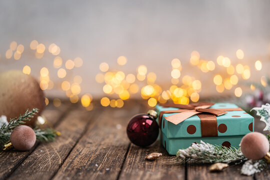 A Christmas background featuring a chic teal polka dot gift box tied with a brown satin bow beside a blush velvet ornament and frosted pine on rustic wood, lit by warm twinkling lights.