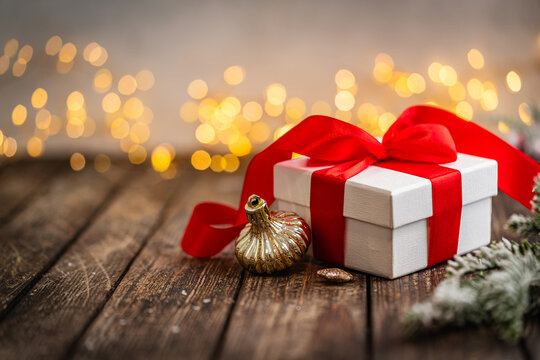 A Christmas background featuring a classic white gift box tied with a bright red satin bow beside a golden ornament and frosted pine on rustic wood, warmly lit with twinkling lights.