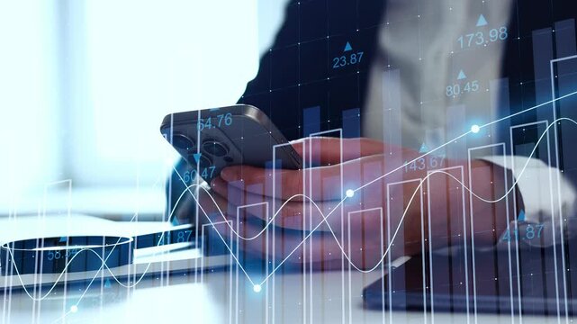 A business person analyzes financial data on a smartphone while seated at a desk. Charts and graphs are visible in the background, showing market trends. The setting is likely an office.