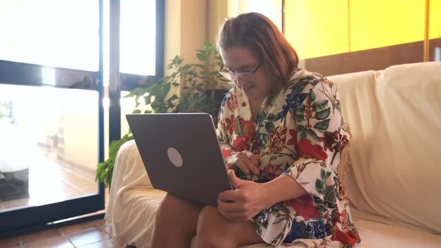 Lockdown shot of adult woman with Down syndrome using laptop while working on project sitting on couch in living room at home during daytime