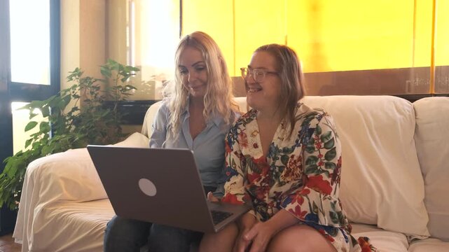 Slow motion shot of happy mature woman with Down syndrome and female friend waving at laptop during video call sitting on sofa in living room at home