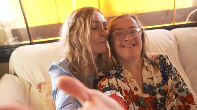 Adorable daughter with Down syndrome and her loving mother taking a happy selfie on a sofa. Loving family moments celebrating inclusion