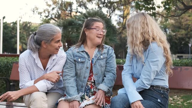 Three happy women, a mother and her two daughters, one with down syndrome, enjoying a pleasant conversation while sitting together on a bench