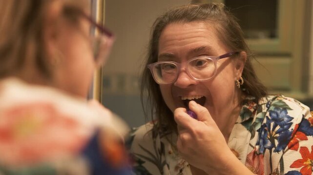Slow motion shot of mature woman with Down syndrome in eyeglasses applying lipstick in front of mirror at home