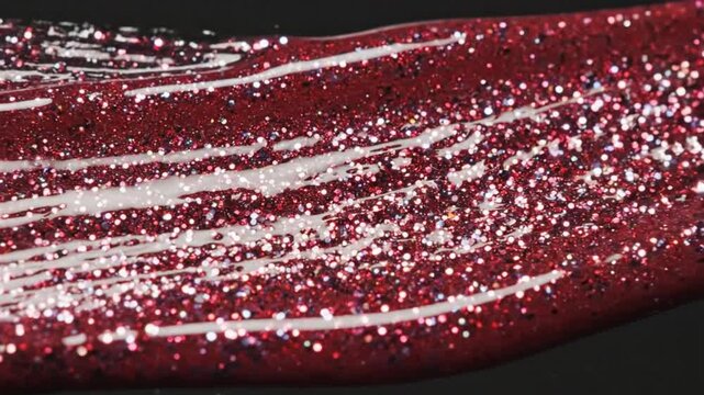 Close-up of sparkling red glitter nail polish being applied with a clear applicator, showing texture and light reflection