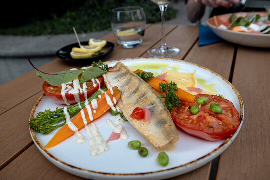 Wroclaw, Poland - July 10, 2026: Restaurant table setting features a plated fish fillet with tomato, vegetables, herbs and sauce at an outdoor meal.