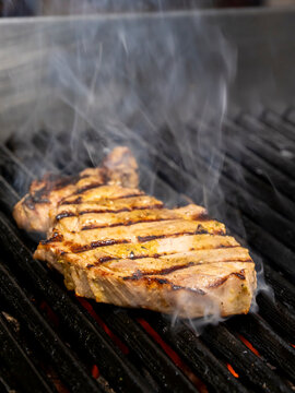 Close-up of a thick pork bondiola steak grilling over hot charcoal. The meat has visible grill marks, a golden-brown surface, and rising smoke, creating an appetizing barbecue cooking scene.