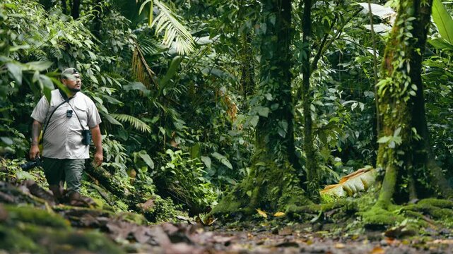 Courageous latino man with a camera and binoculars exploring a lush green rainforest in Costa Rica, hiking on a muddy trail surrounded by trees