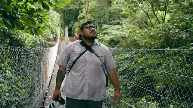 Hispanic male photographer hiking on a suspended bridge. Tourist exploring the lush green rainforest in Arenal Volcano National Park