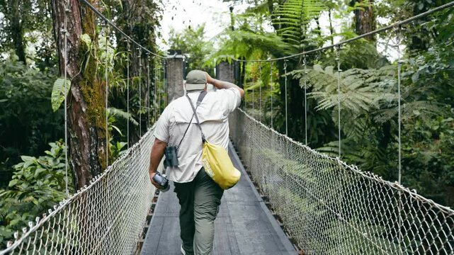 Latino explorer with a camera and binoculars walking across a hanging bridge. Lush green jungle in the Arenal Volcano National Park, Costa Rica