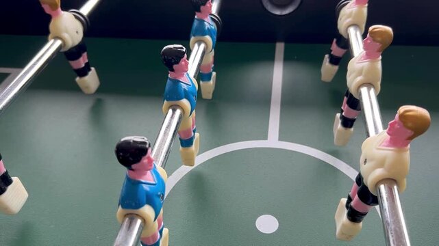 Camera moves across a foosball table, showing miniature players mounted on metal rods over a green playing field