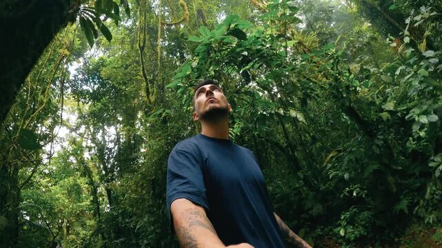 Young man with tattoos filming a selfie video while walking through a lush tropical rainforest, looking up at the canopy in amazement