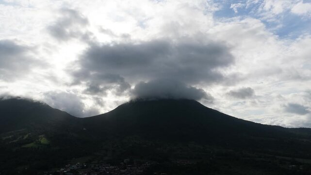 Majestic Arenal Volcano silhouetted against a bright sky. Dark, dramatic clouds floating over its peak with the town of La Fortuna below