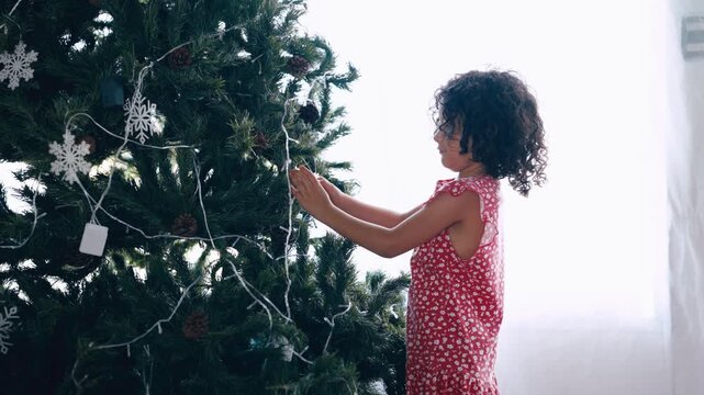 Happy little girl with curly hair carefully places ornaments on a christmas tree. Festive holiday preparation and family tradition concept