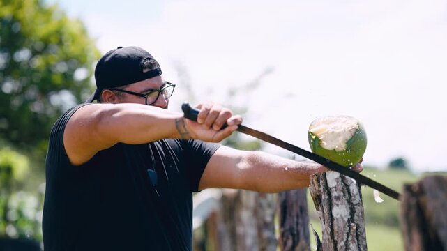 Focused man wearing glasses and a cap skillfully using a large knife to open a fresh green coconut on a wooden post in the countryside