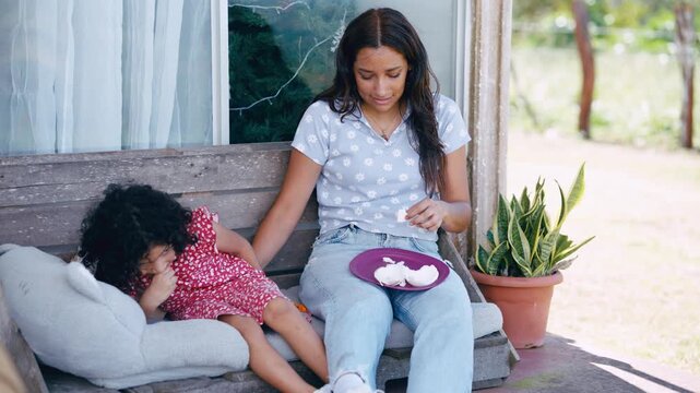 Sad mother sitting on a porch bench next to her little daughter. The woman eats something from a plate while wiping away tears