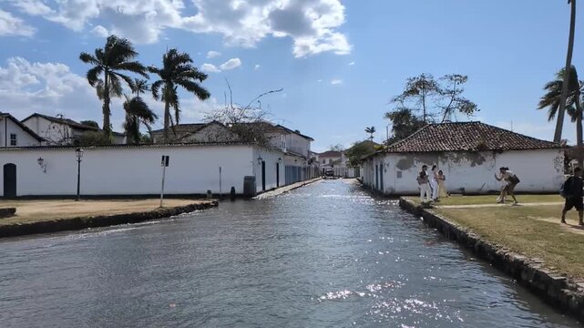 Static slow motion shot of high tide sea water flooding cobblestone street in Paraty, Brazil, with tourists and palm trees on a sunny day.