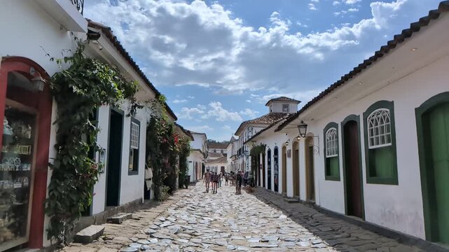 Static slow motion footage of tourists walking down a cobblestone street lined with historic colonial buildings in Paraty, Brazil, under a cloudy blue sky.