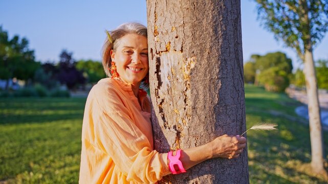 Mature woman smiling, embracing a tree trunk in a park, holding a blade of grass