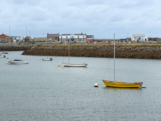 A bright yellow sailboat rests on calm water in Howth Harbour, with additional small boats and the working waterfront beneath an overcast sky. © Ryan