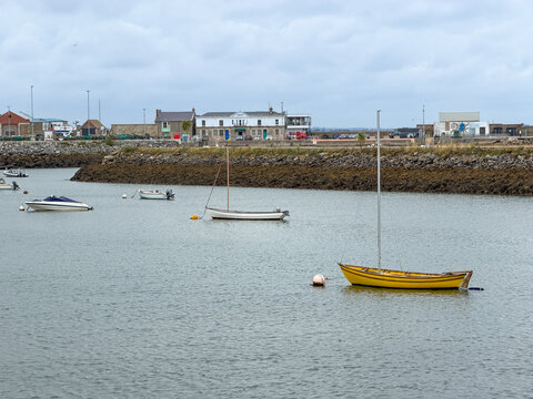A bright yellow sailboat rests on calm water in Howth Harbour, with additional small boats and the working waterfront beneath an overcast sky.