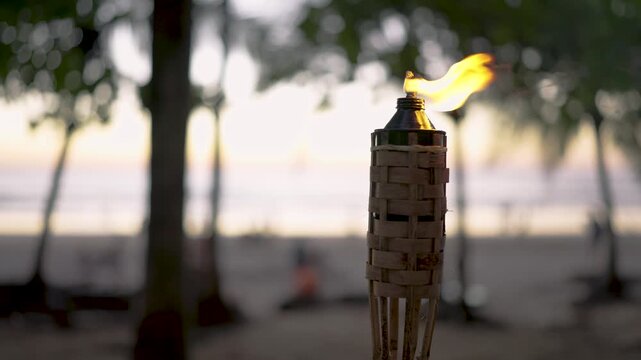 Close up of a burning tiki torch illuminating a tropical sandy beach with palm trees. The warm flame flickers gently in the evening breeze