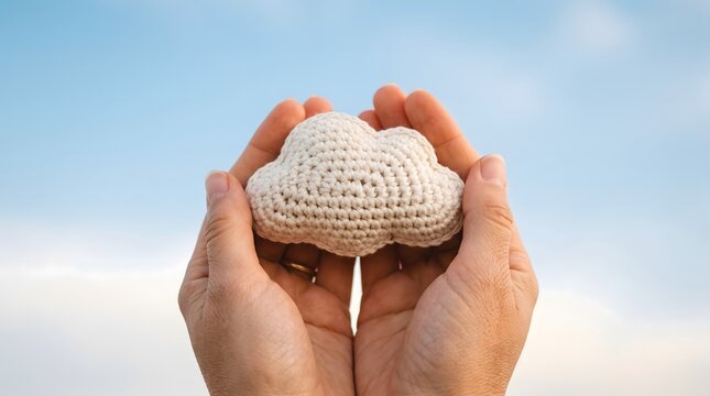 Hands holding a small white crocheted cloud toy against a bright blue sky with sunlight