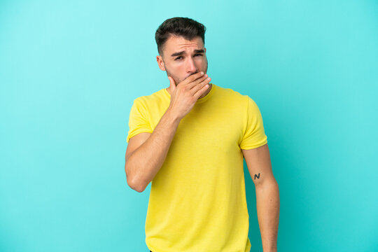 Young caucasian man isolated on blue background yawning and covering wide open mouth with hand