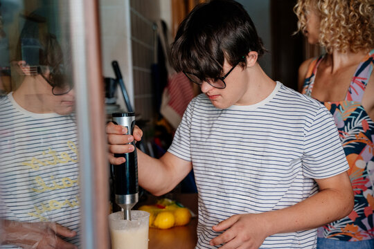 Teenage boy with Down syndrome using hand blender while preparing food with mother in home kitchen