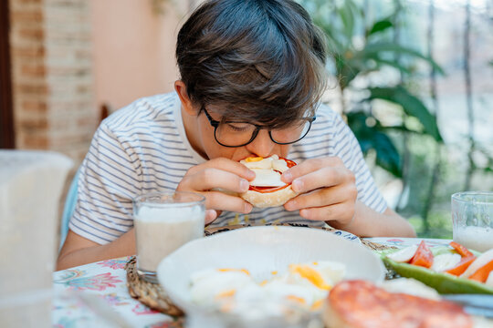 Teenage boy with Down syndrome eating homemade breakfast sandwich outdoors on family terrace