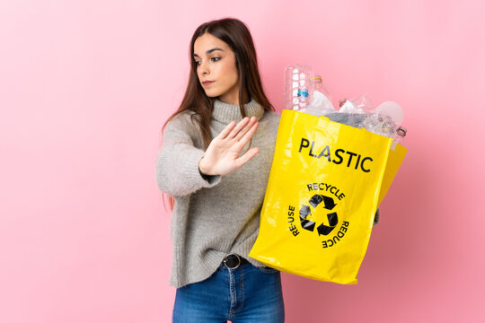 Young caucasian woman holding a bag full of plastic bottles to recycle isolated on pink background making stop gesture and disappointed