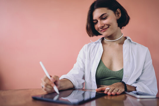 Smart tech integration cozy productivity lets cheerful Caucasian female freelancer work on tablet screen with stylus pen at wooden table, promoting hybrid lifestyle personal autonomy