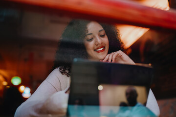 Smiling young woman with curly hair looking down at smart tablet device screen behind cozy cafe window glass reflection, creative freelancer working remotely or student e-learning online © BullRun