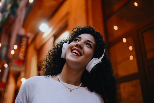 Close up portrait of happy young woman listening to music in wireless headphones while walking on vintage evening street, cheerful female tourist enjoying audio guide or online podcast